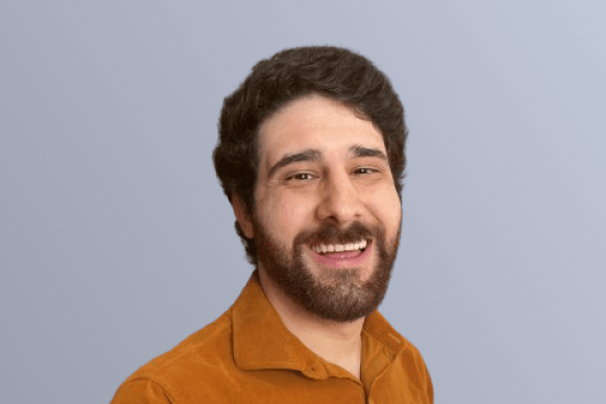 Smiling man with dark hair and beard wearing an orange shirt, posing against a plain light blue background.