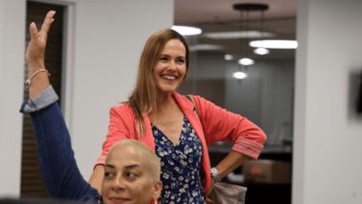 "Two women smiling in an office, one raising her hand and the other standing with a hand on her hip."
