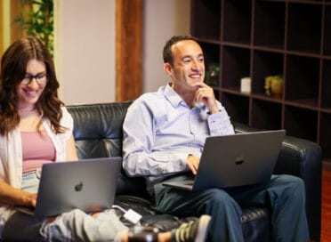 Two colleagues sitting on a couch with laptops, smiling and engaged in conversation in a modern office setting.