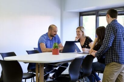 Group of four people collaborating around a conference table with laptops and a small potted plant in an office setting