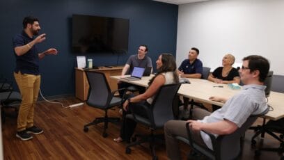 A man gives a presentation to a group of five people seated around a conference table in an office.