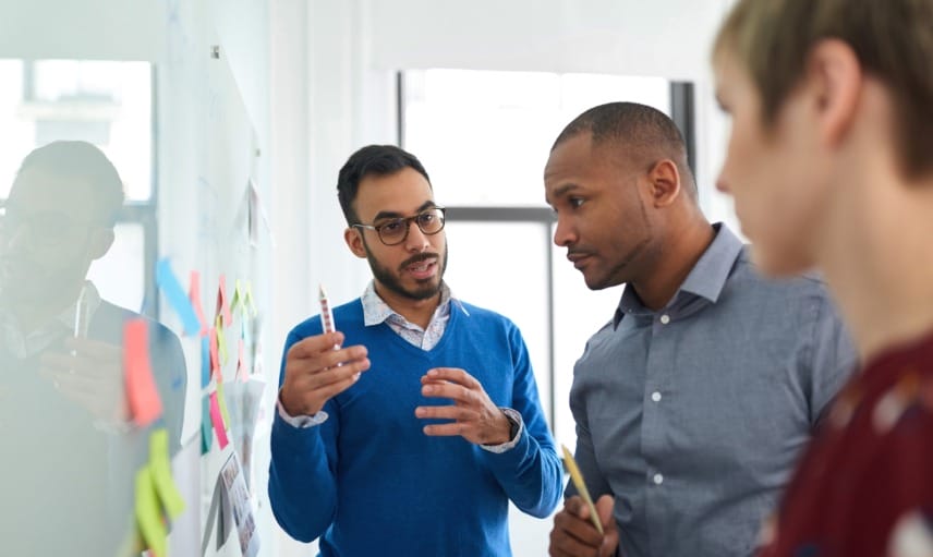 Three colleagues review sticky notes on a glass whiteboard, aligning plans and discussing next steps.